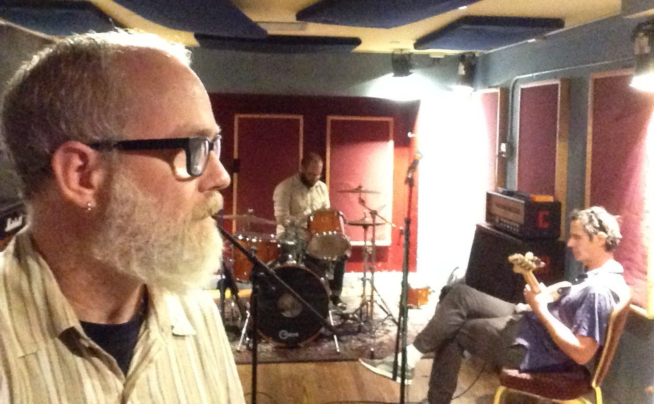 A gray-bearded man is seen in profile at the left side of the image, while behind him in a small rehearsal studio sit a bearded man playing drums and clean-shaven man playing a bass guitar in his lap. Microphones and amps are also visible.