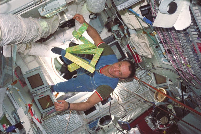 A cosmonaut floats inside the space station Mir, his body rotated 90 degrees clockwise. In front of him floats the Cosmic Dancer sculpture, which looks a bit like an angular, headless snake, its body bent at random angles and its sides squared off rather than rounded. The sculpture is light green with multi-colored streaks, and about 1.5 feet across in every direction.