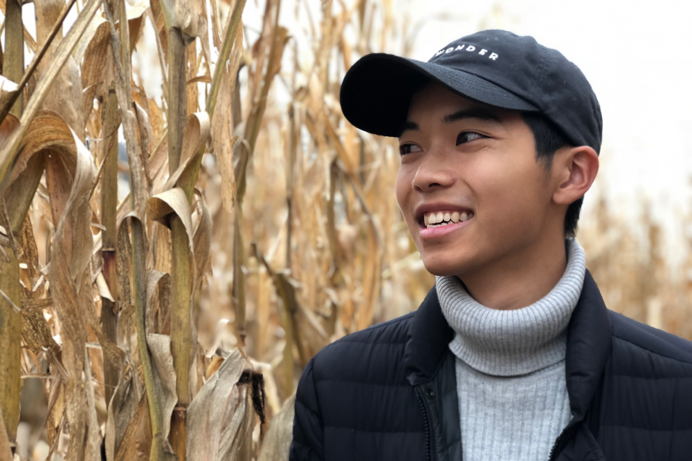 Jason in a black cap and grey turtleneck and black jacket smiling and looking to the right in front of a corn maze background.