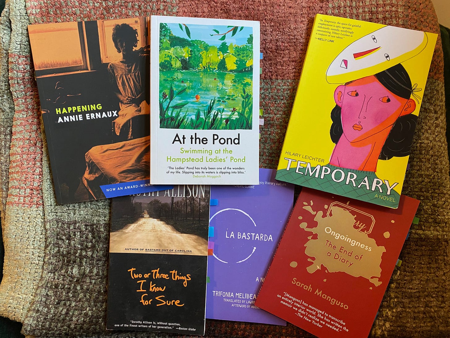 Six short books spread out on a red and green blanket: Happening, At the Pond, Temporary, Two or Three Things I Know for Sure, La Bastarda, Ongoingness.