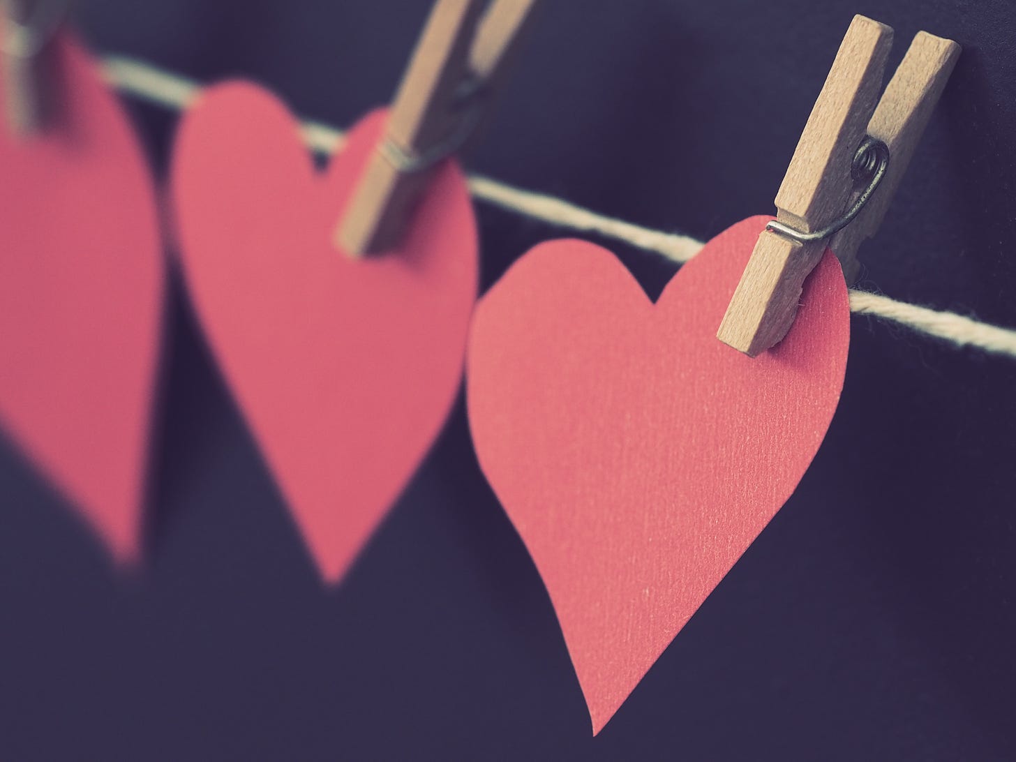 Three red paper hearts are hanging on a twine string, and are attached by clothes pins. It's against a black background and like, it really pops.