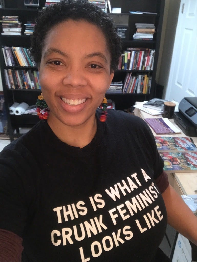 This is a headshot image of a light Brown Black woman with thick eye brows, high cheek bones, full lips, short black curly hair and she is smiling. She has multi-colored wood hoop earrings, a black tshirt with white capital letters that spell This is What A Crunk Feminist Looks Like. There is a black bookcase with stacks of books in the background.