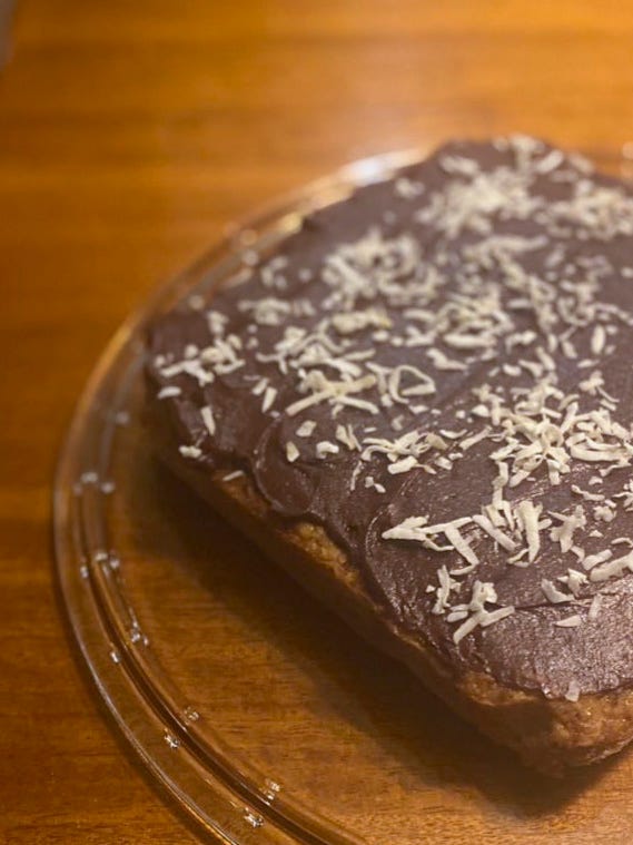 A square cake on a glass plate, with chocolate icing spread on top and shredded flakes of coconut sprinkled over.