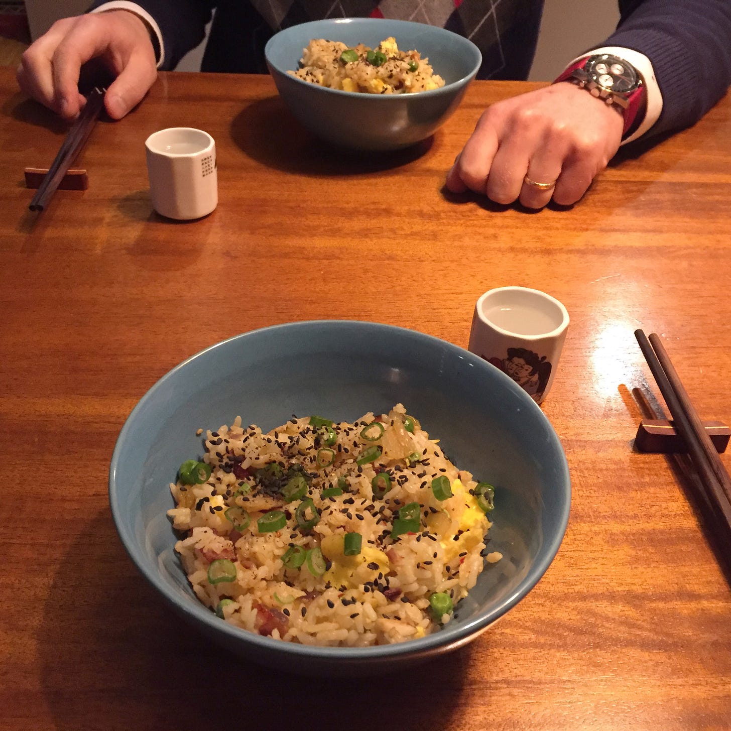 Two blue bowls of fried rice topped with black sesame seeds and green onions. Next to the bowls are wooden chopsticks and two small white cups of soju.