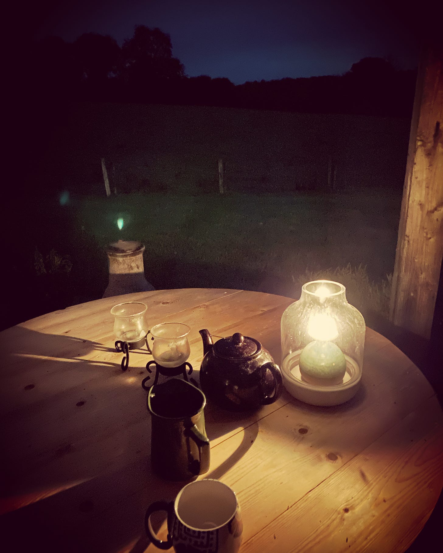 A small table holds a tea kettle and mug, and is lit by candlelight in the night.