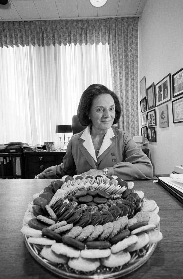Frances Hesselbein in a black-and-white photo with her hair swept back and seated at a table in front of a platter of cookies.