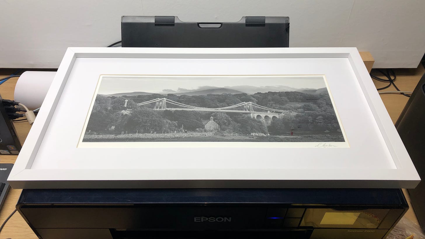 White frame and matte board around a panormic monochrome photographic print showing a church, bridge and mountains behind.