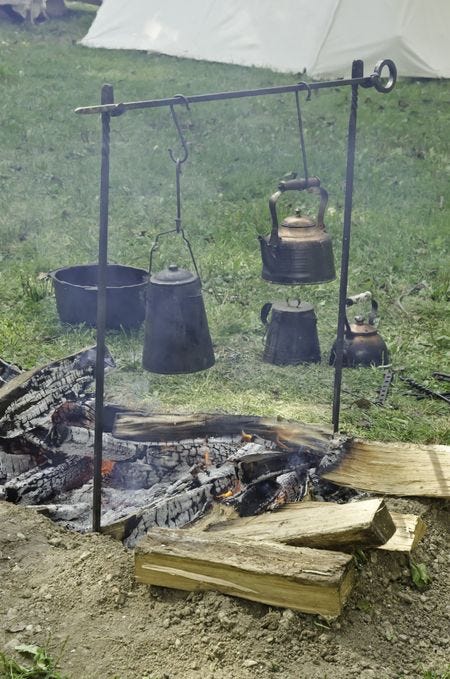 A field of green grass, with a canvas tent in the baclground at the top. In the foreground is a smoking campfire set up with two kettles, more kettles and a pot just beyond, with extra wood nearby.
