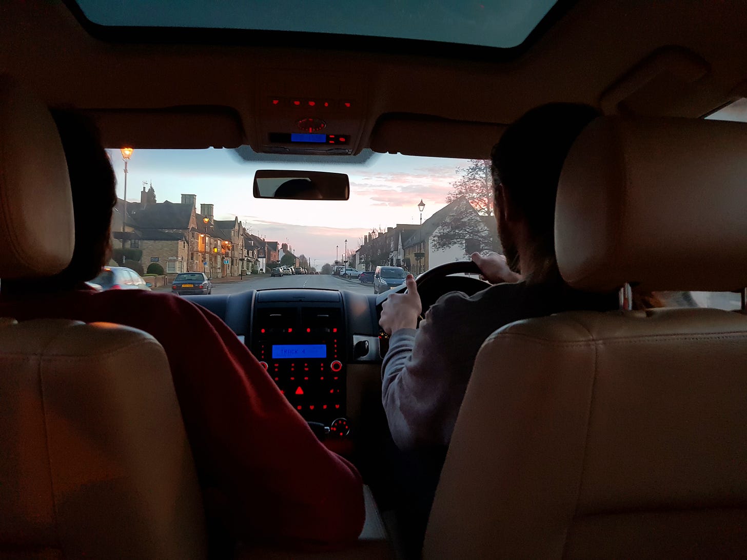 Photo taken from the back seat of a car shows a young man driving and an older man in the front passenger’s seat. Through the windshield, a sunset can be partially seen lighting the sky over a village high street, with shades of pink and orange.
