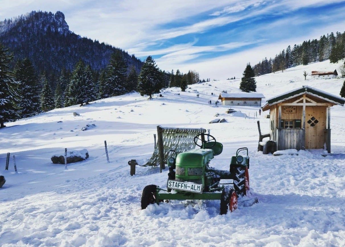 A snowy mountainside with forests behind. In the foreground is an old green tractor with a number plate that says "Staffn-Alm". there is a small wooden hut behind it.
