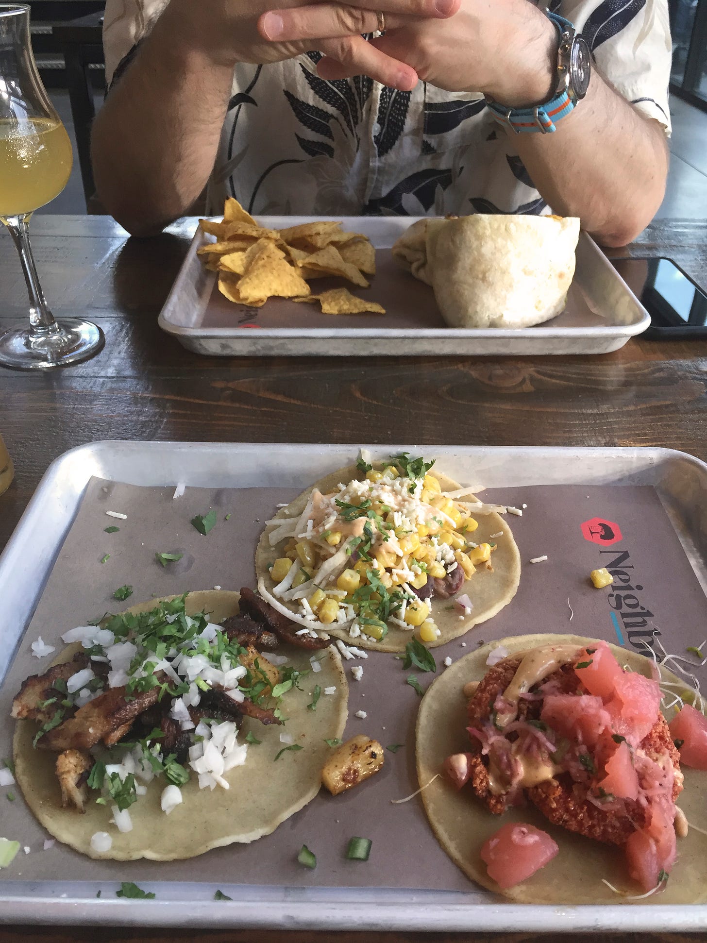 On a metal tray lined with paper are three different tacos, sprinkled with cilantro. Across the table on another tray is a burrito cut in half and a small pile of yellow corn tortilla chips. Jeff's hands are linked together overtop of the tray as his elbows rest on the table.