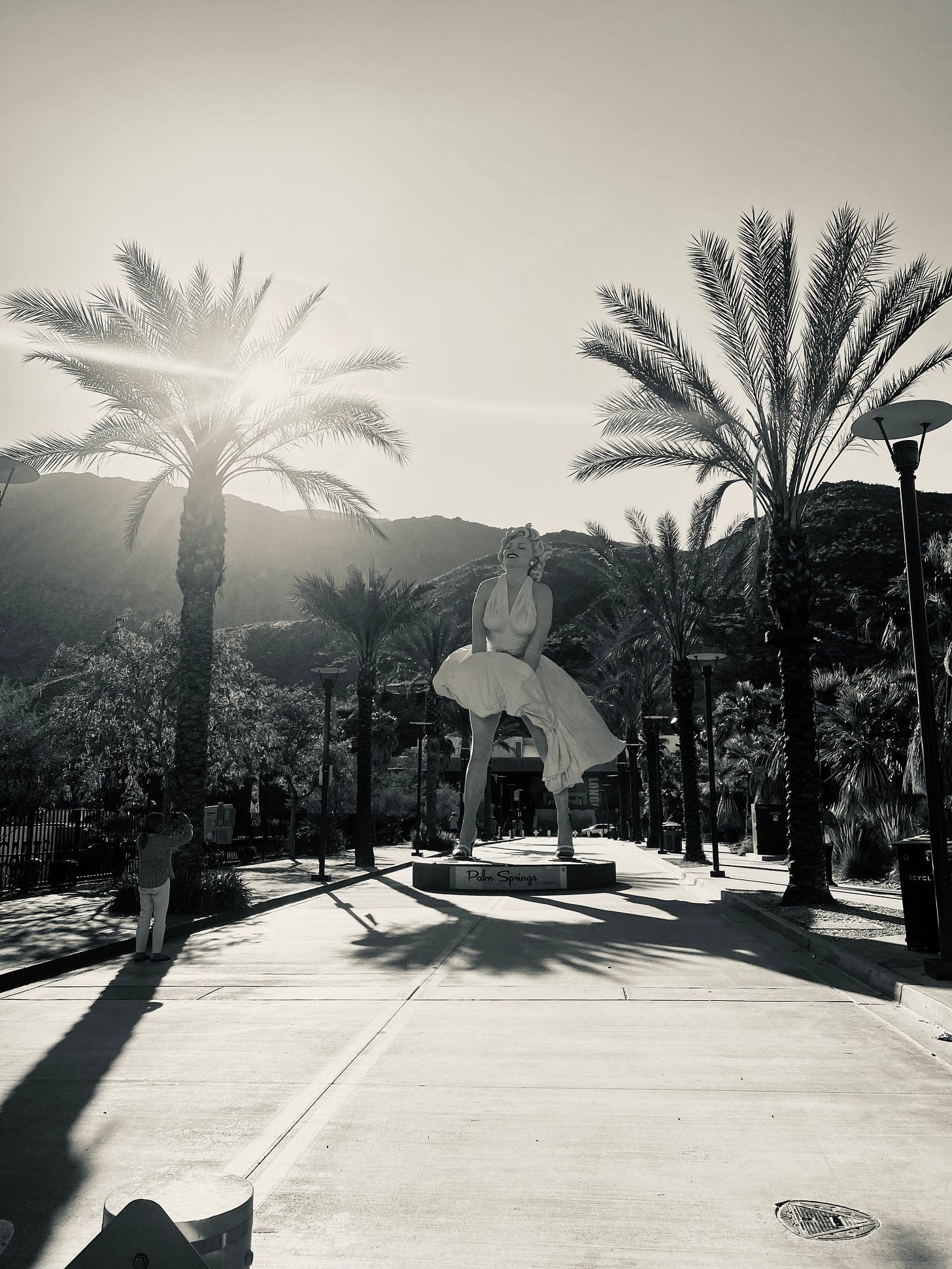 Larger than life Marilyn Monroe statute in Palm Springs, the author’s mother taking a photo off to Marilyn’s right; late sun light peaking through the palm fronds.