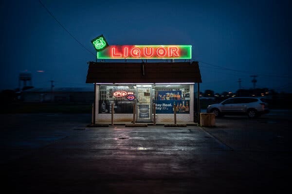 A small liquor store, with the word “liquor” in bright neon lights on the roof, sits in the middle of a parking lot at dusk.