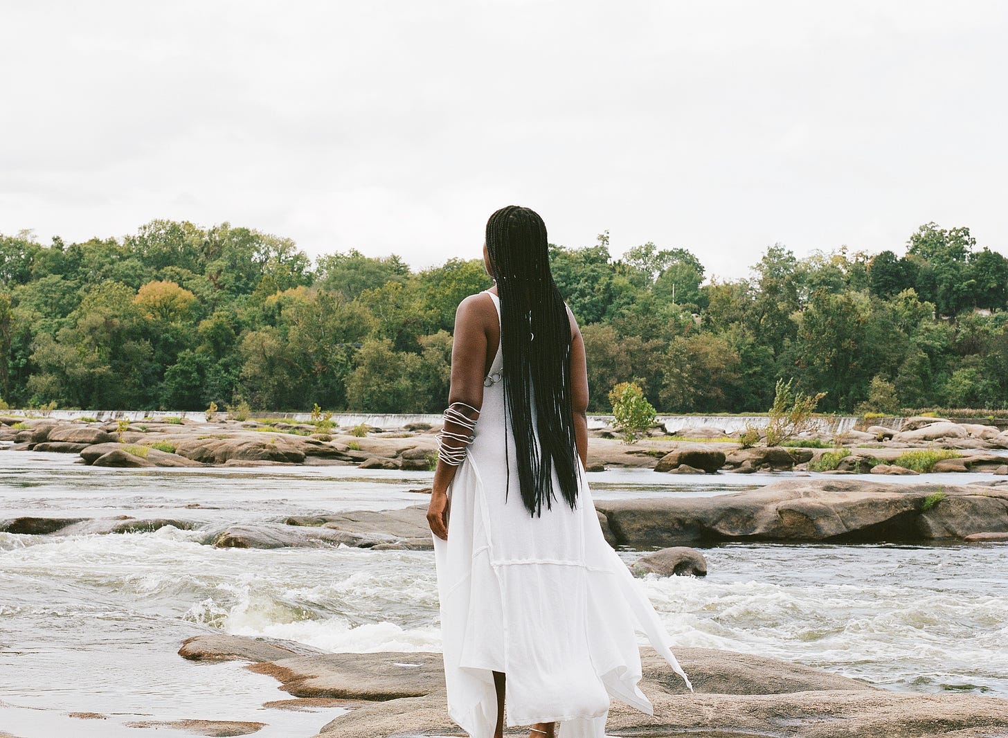 A black woman (me) with braids wearing a white dress, back facing the camera while standing on top of river rocks with a tree line in the background.