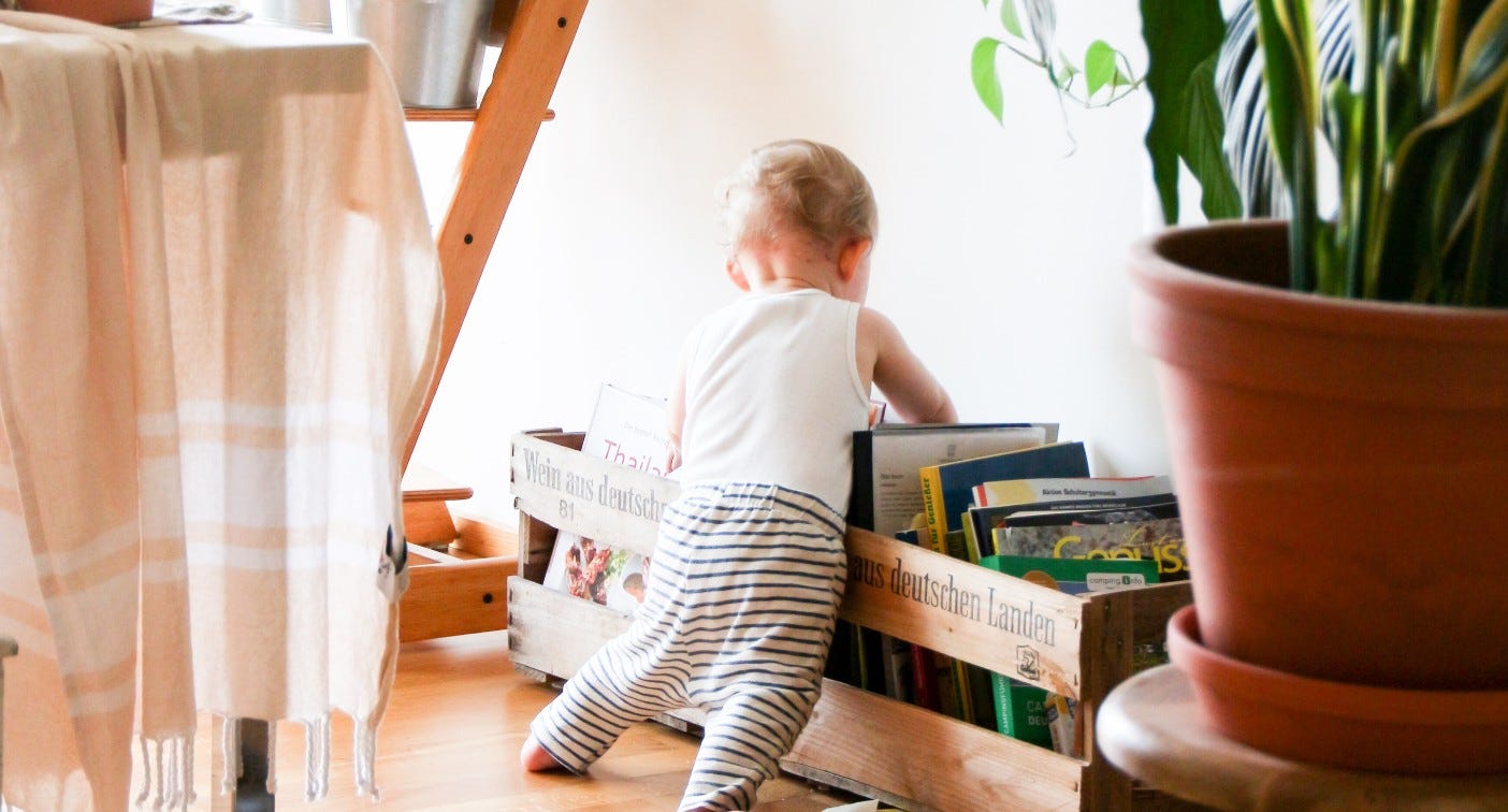 A photo of baby from behind. The baby has pulled itself up to look at a crate filled with books. (Photo by Brina Blum on Unsplash)
