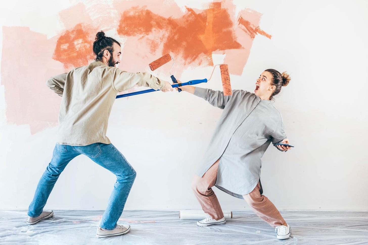 A man playfully stretches a paint roller towards his partner, who looks suprised.