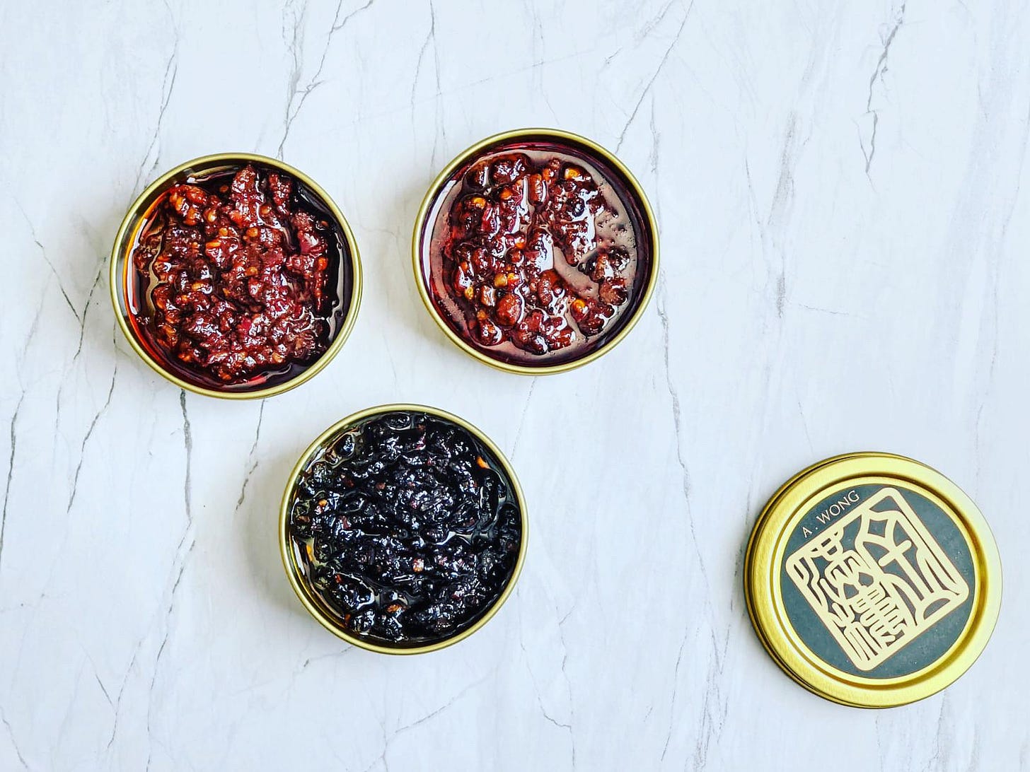 An overhead shot of three open tins of sauces and a tin lid on a marble tabletop