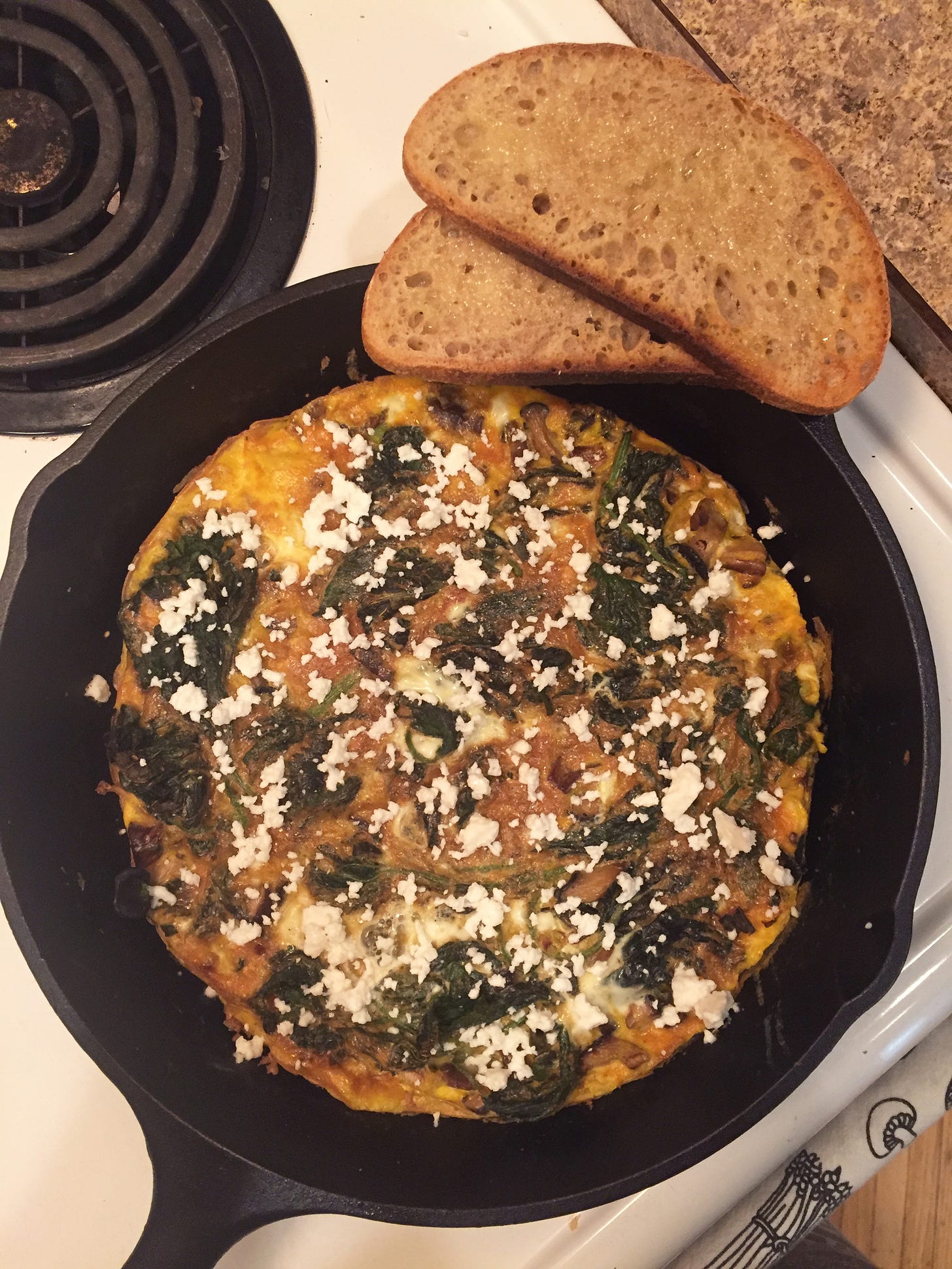 A cast iron pan sits on the stove, filled with a frittata of mixed mushrooms and dark green nettles. Crumbles of feta across the top contrast the deep yellow of the egg, and resting at the edge of the pan are two slices of whole wheat sourdough.