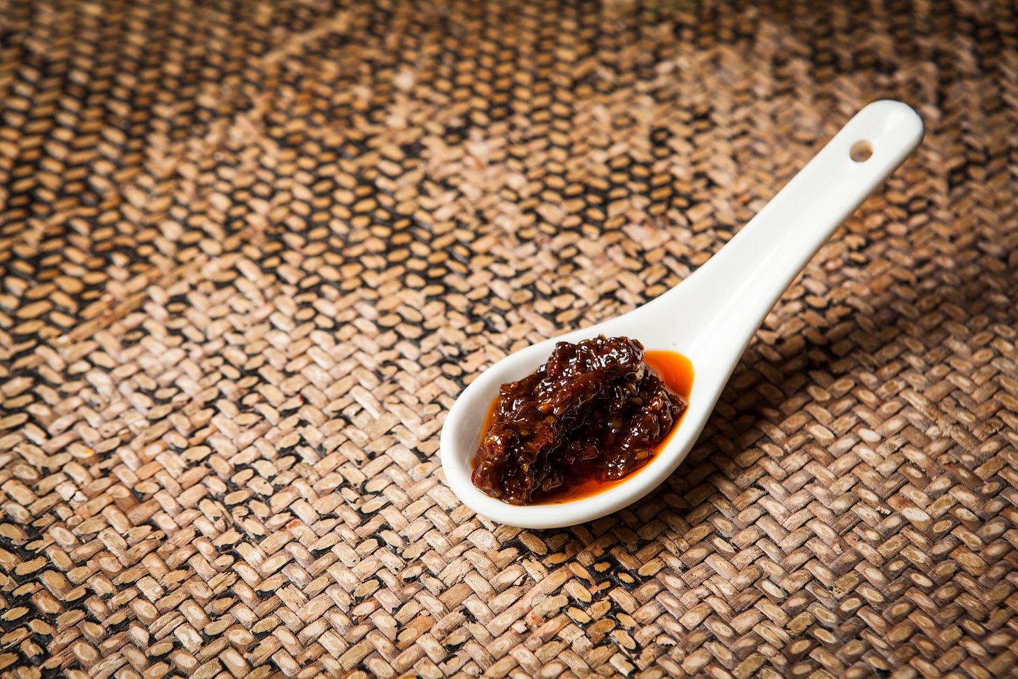 A white ceramic spoon rests on a rattan table mat. In the bowl of the spoon is a deep-reddish brown relish.