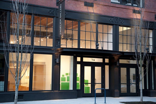 The outiside of an art gallery housed in a brick building with black steel beams. The lights are on in the gallery, causing a warm glow from inside. A sign hung to the left of the door reads "Froelick Gallery."