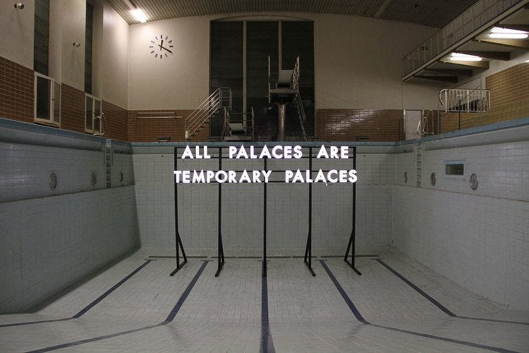 An empty swimming pool is half-lit, with a tall iron rack rising from the floor of the empty basin. On it, lit-up letters spell out ALL PALACES ARE TEMPORARY PALACES