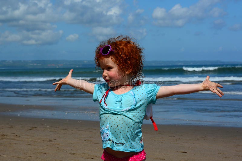 Little girl with curly red hair and blue t-shirt standing on beach with arms wide open and the ocean behind her
