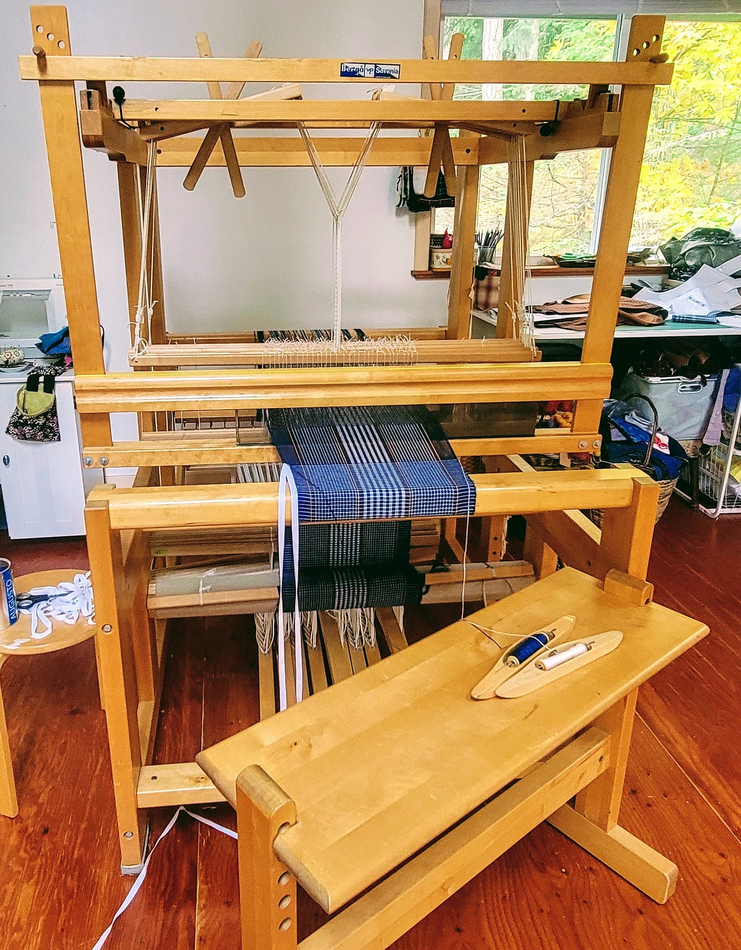 A weaving loom with some weaving on it, messy table in the background. 