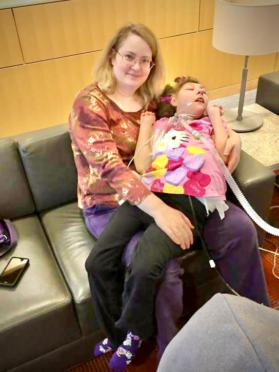 A blond white woman with glasses smiles at the camera while sitting with a teenage girl on her lap. The girl has a bright pink Hello Kitty shirt on as well as a tracheostomy tube and a g-tube. 