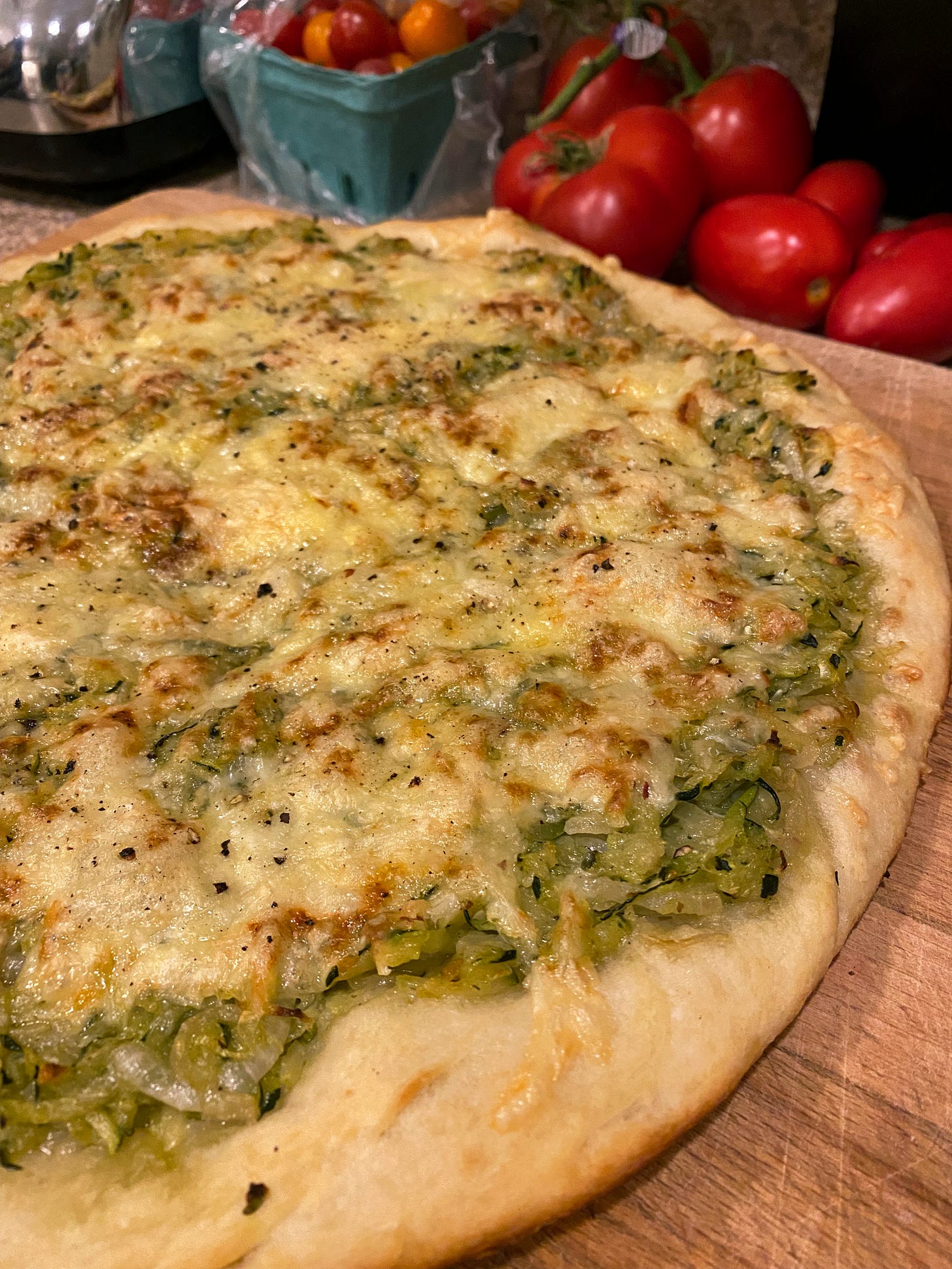 On a wooden cutting board, an unsliced pizza covered in shredded zucchini and melted, browned cheese. 