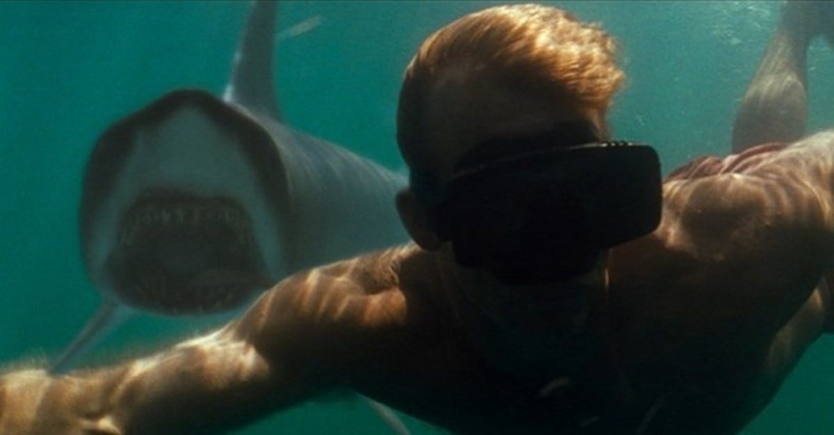 A man swimming underwater towards the camera. Over his shoulder is a shark with its mouth open turning towards him.