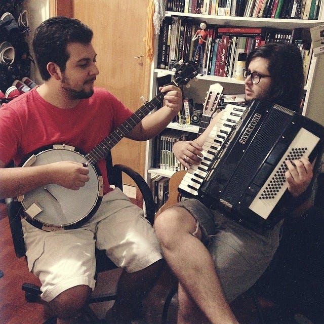 Angelo playing the accordion with his friend, Tiago, playing the banjo. Behind them, a bookshelf full with books.