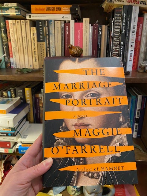 Hand holding hardcover of The Marriage Portrait in front of a bookcase