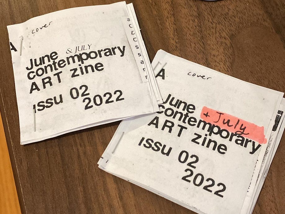 Two black-and-white zines titled "A June & July Contemporary Art Zine," set against the backdrop of a wooden table. The zines are stapled together.