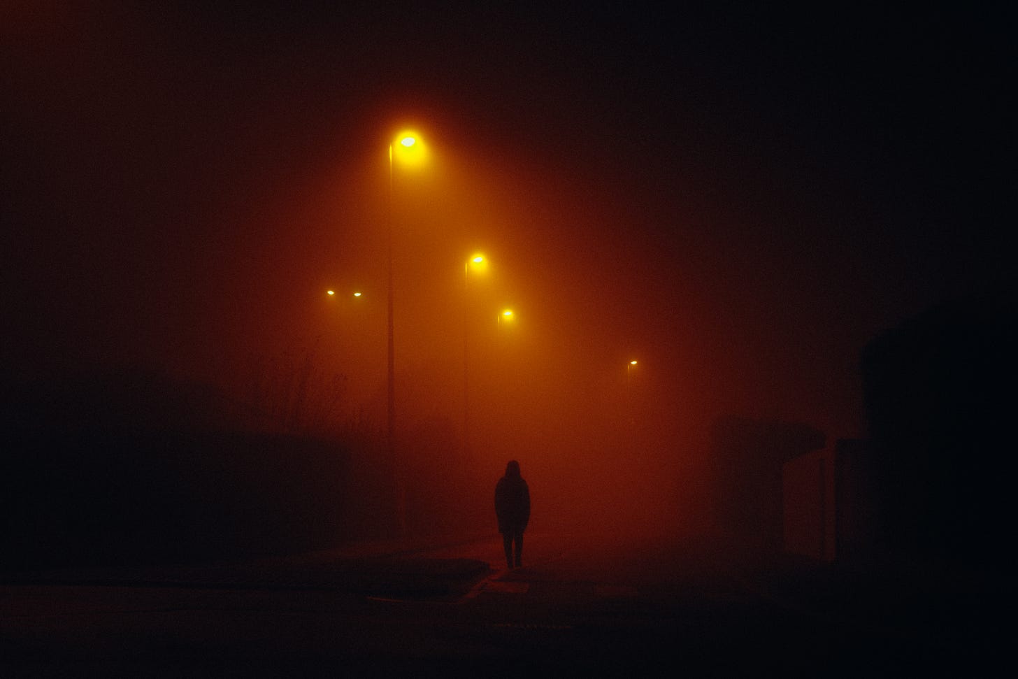 A lone human figure walks down a street under lights in a rust colored haze