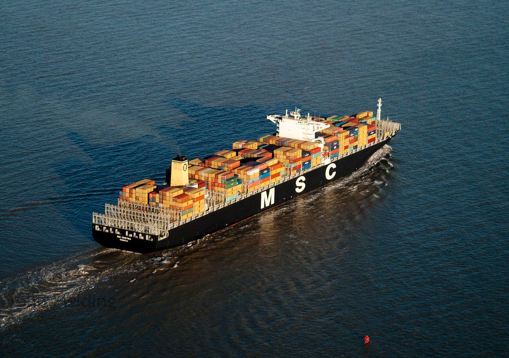 A ship with a lot of containers on it and MSC on the side sails in calm waters. the smoke stack at the back is billowing smoke, while the bridge pokes out from among the containers full of consumerist rubbish