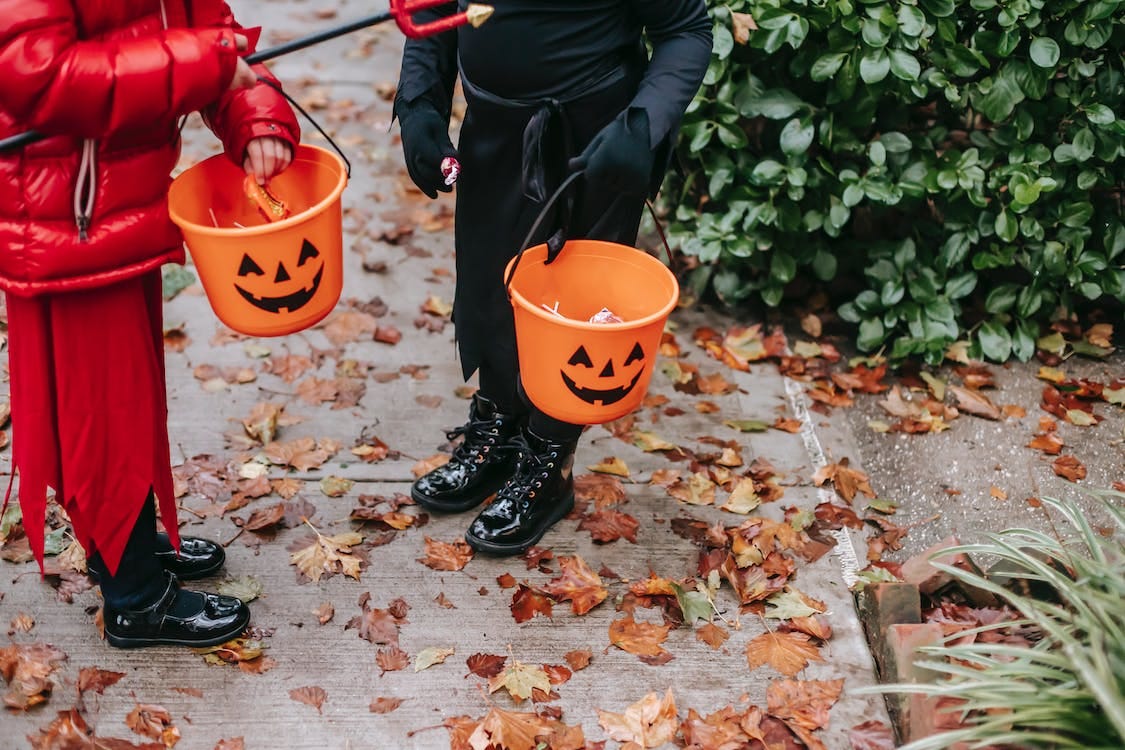 Free Crop unrecognizable kids in red devil costume with pitchfork and in black witch costume on Halloween with buckets full of candies standing on road in autumn day Stock Photo