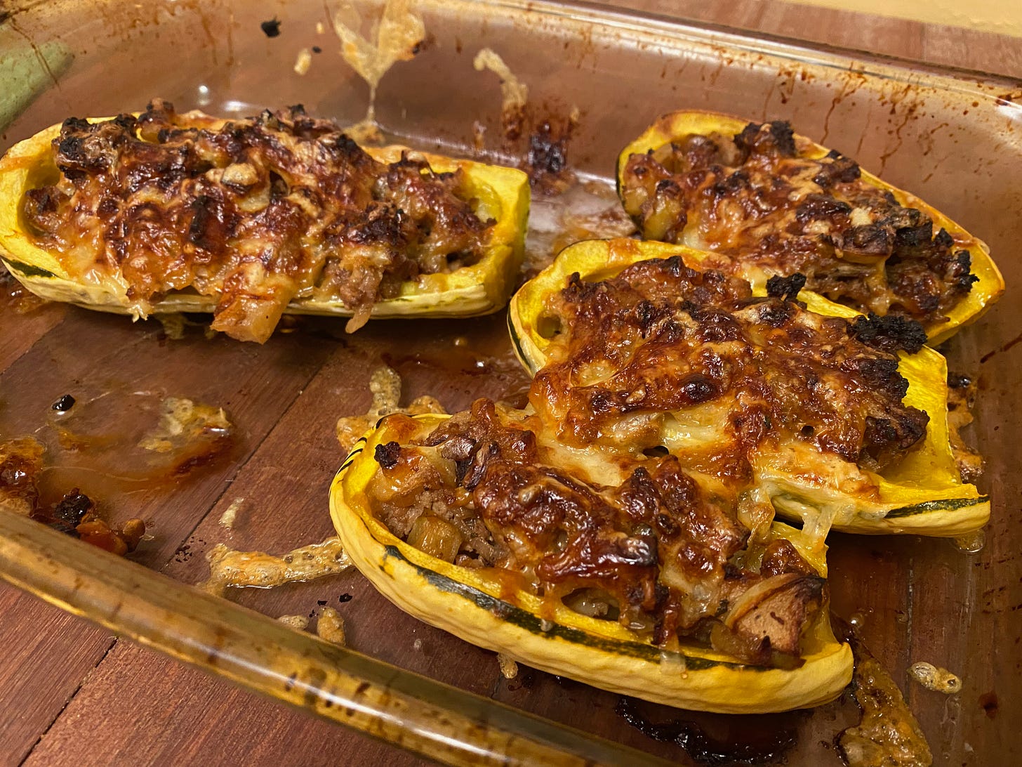 A glass baking dish holds four halves of delicata squash, overflowing with filling and topped browned and bubbly cheese. There is an empty space in the front of the pan where a fifth half used to be.