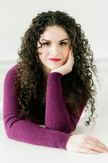 A white woman in her mid-twenties with long, very curly brown hair, stares into the camera. She is wearing a purple v-neck sweater and red lipstick. Photo credit: Astrid Photography