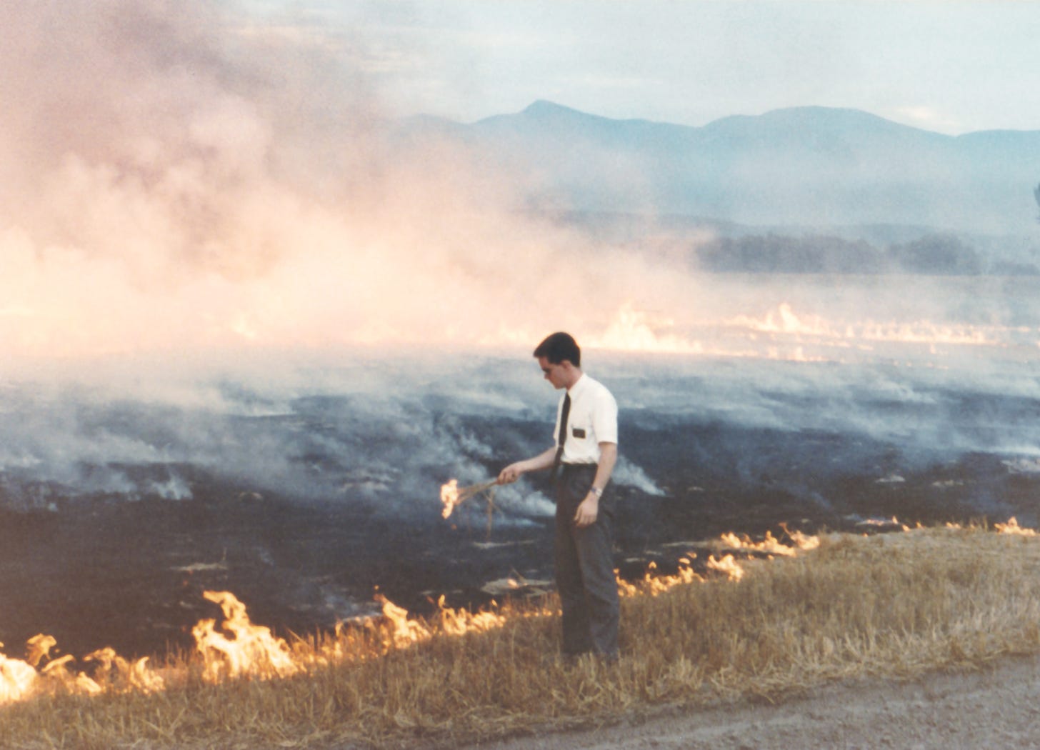 A Mormon missionary wearing dress clothes and a black name tag, stands before a field of burning, smoking wheat stubble holding a sheaf of flaming wheat.