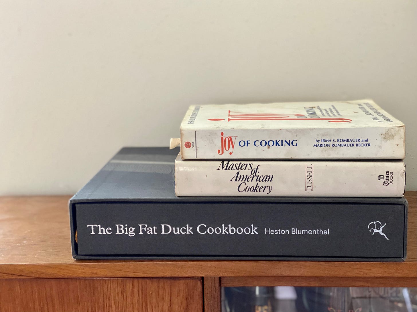 A stack of three books on a wooden shelf: The Joy of Cooking, Masters of American Cookery by Betty Fussell, and The Big Fat Duck Cookbook by Heston Blumenthal