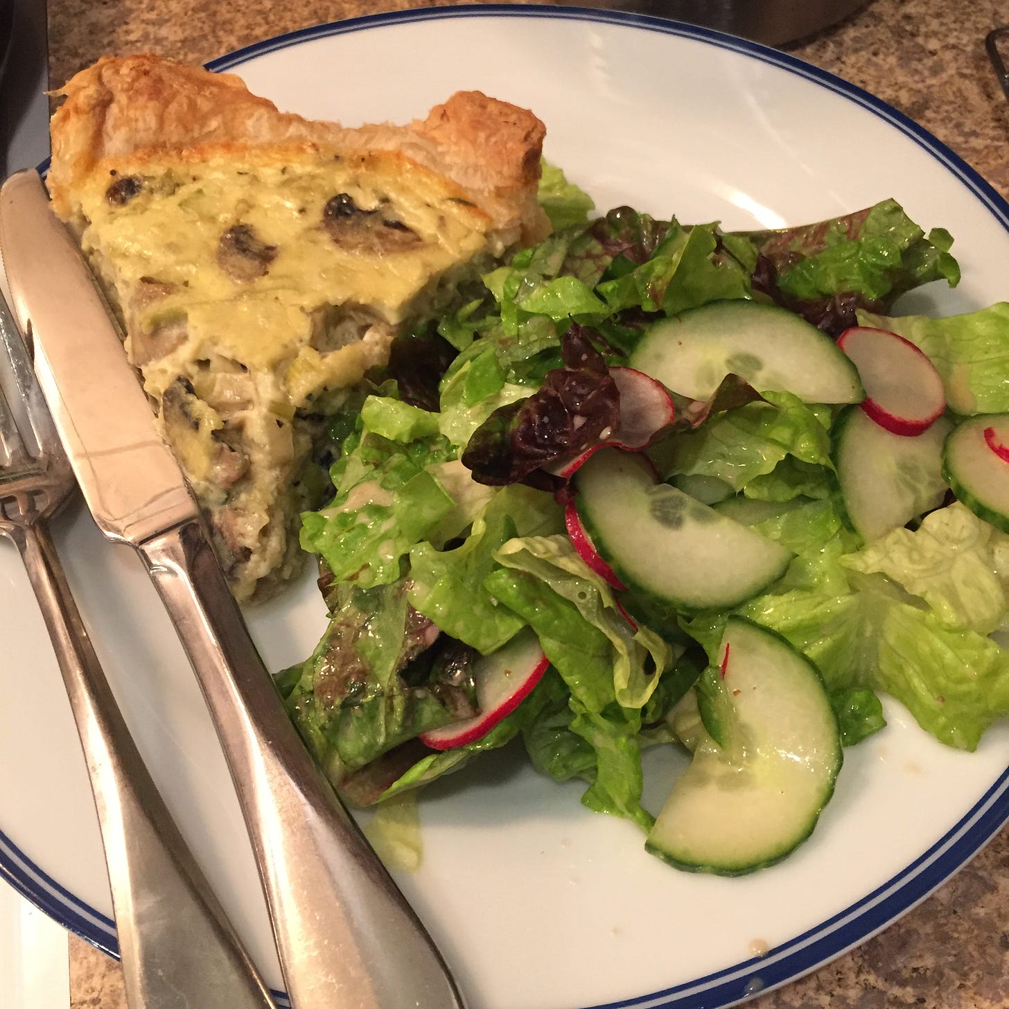 On a white plate with a blue rim, a triangle of quiche with crisp brown edges and pieces of mushrooms and leeks throughout. On its left are a knife and fork, and on its right is a large portion of green salad with cucumber and radish.