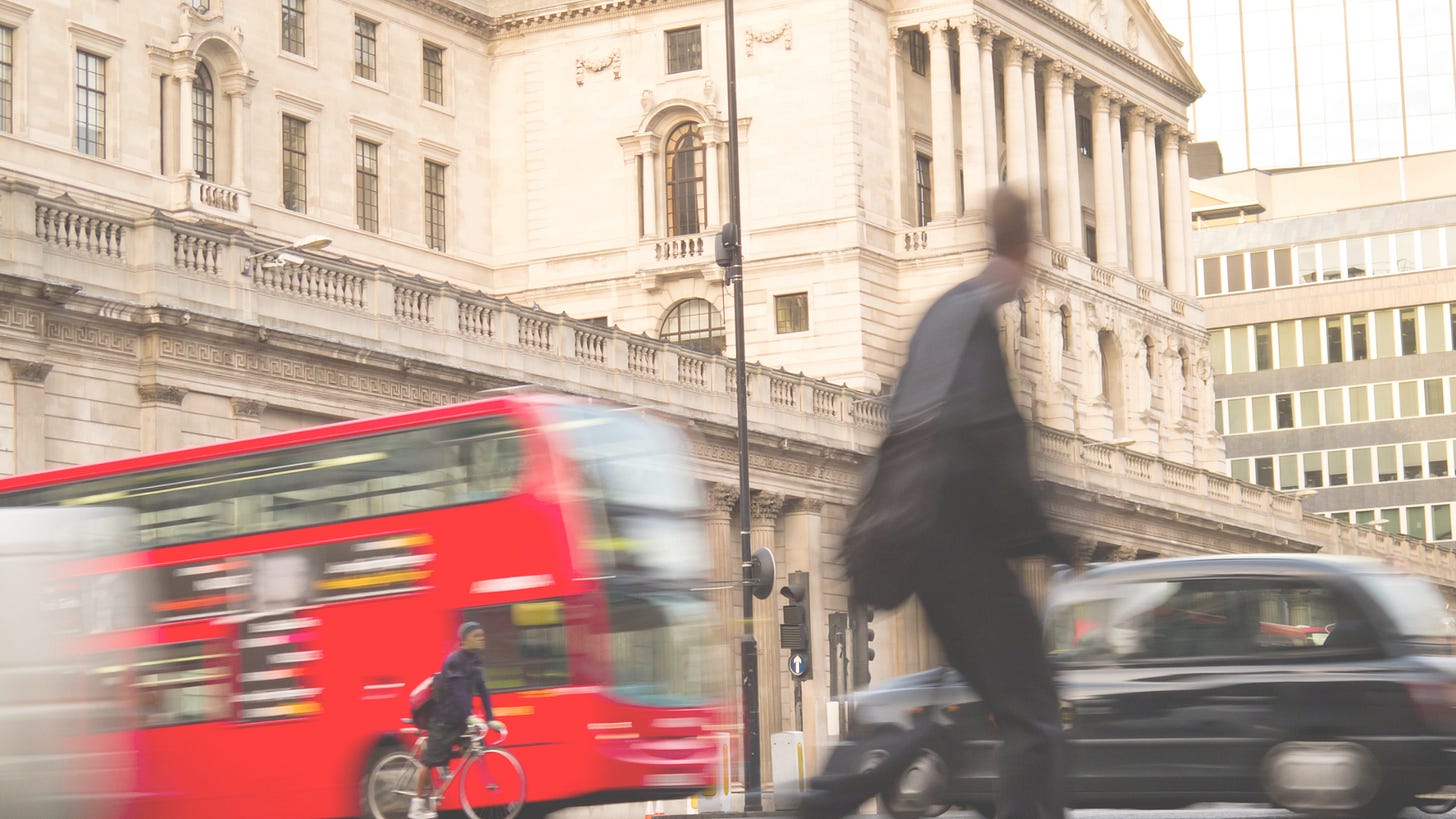 blurred image of a red double decker bus, a man riding a bicycle, a pedestrian, and a black car with an old building in the background