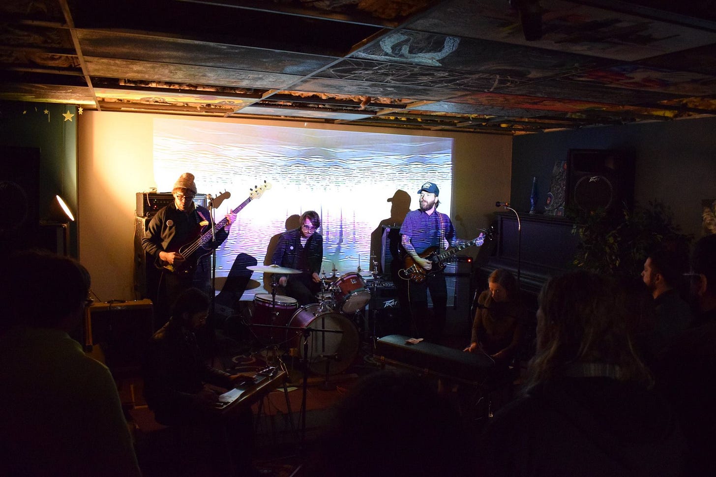 A band with two guitars, a drummer, a keyboard player, and a mandolin player perform on a small, dark stage. A projector screen provides a colorful ripple effect in the background. A band with two guitars, a drummer, a keyboard player, and a mandolin player perform on a small, dark stage. A projector screen provides a colorful ripple effect in the background.