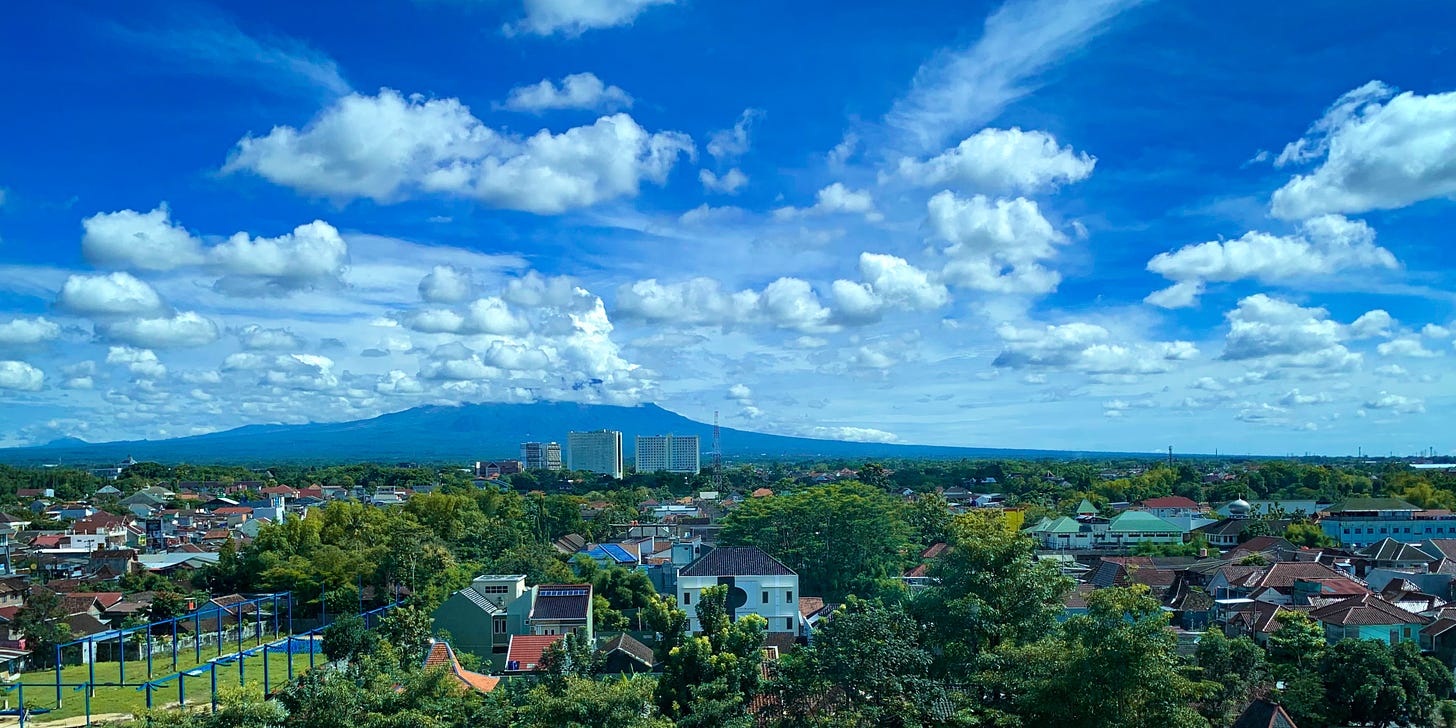A landscape view of a mountain, its top hidden in the clouds. On the foot of the mountain are houses and trees. There's a couple of tall buildings ruining the view.