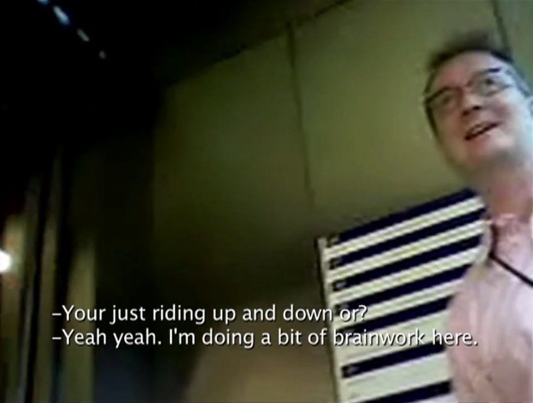 Screenshot of a video. Off frame, the artist secretly recorded the reactions of workers entering and exiting the elevator. The first subtitle reads, you're riding up and down or? The second subtitle reads; yeah yeah, I'm doing a bit of brainwork here.