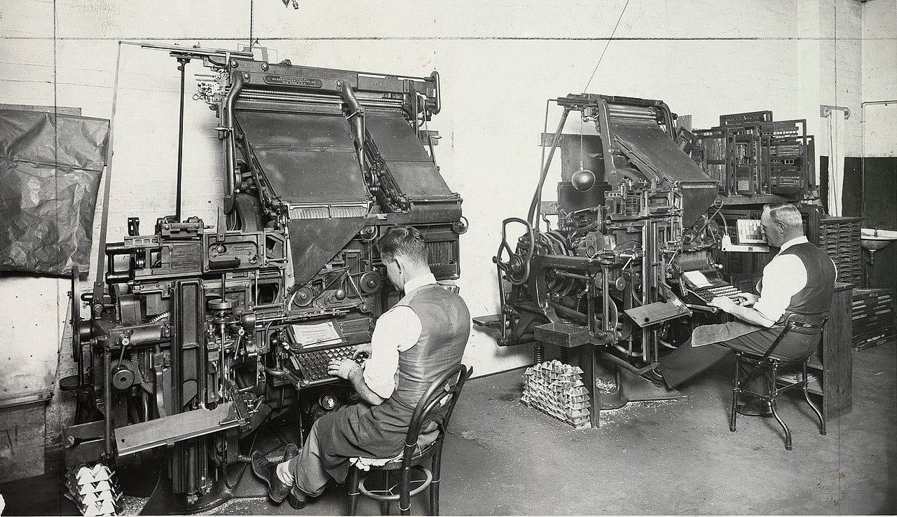 Black-and-white photo of two men seated at linotype machines in a large office. The machines and very tall, made of metal, and have a lot of moving mechanisms.