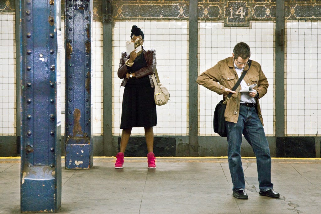 na plataforma de um metrô, uma mulher negra em pé lê um livro brochura; mais à direita, um homem branco lê um folheto