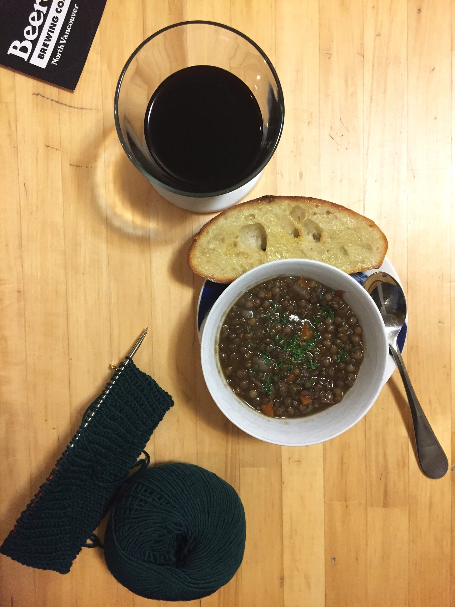 A small bowl of lentil soup topped with parsley sits on top of a plate holding a slice of sourdough and a spoon. Above it is a glass of dark beer, and a black sticker with white text reading "Beere Brewing Co." In the bottom left corner of the photo is a ball of dark green yarn and several rows of knitting on small circular needles.