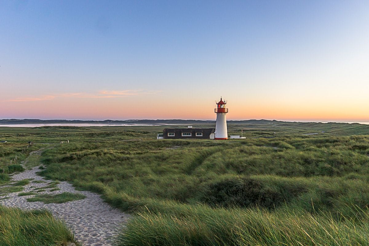 A red and white lighthouse surrounded by long grasses with a beach in the far distance and sunset beyond