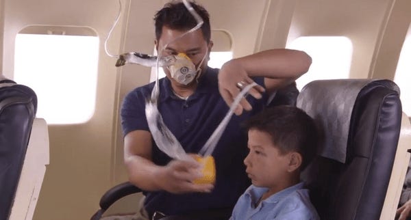 Man with oxygen mask on in an airplane is moving to attach the oxygen mask to the child next to him.